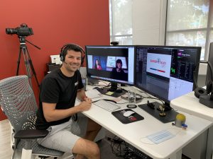 A white man with brown hair sits smiling in front of a two-monitor set up for Badger Talks Live with a red wall in the background