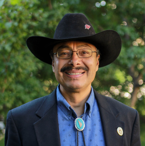 Alfonso Morales' headshot. A man in a cowboy hat with a Wisconsin pin, a sports coat, and a blue adornment on his collar smiles against a green leafy background.