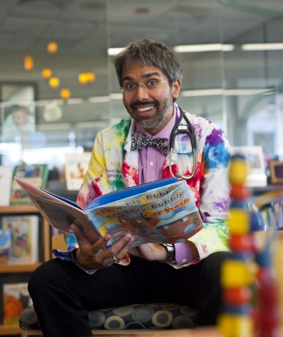 A brown-skinned man with dark hair, wearing a colorful lab coat and stethoscope, smiling and holding an open children's book