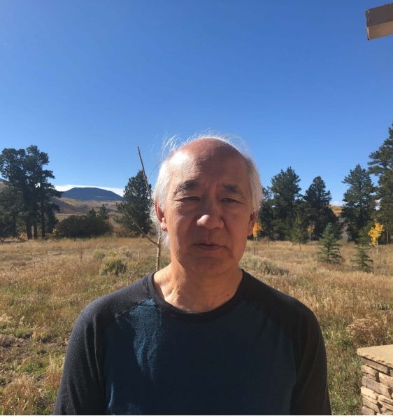 Jerry Yin, a man with white hair and a dark blue shirt, standing in front of a grassy and tree-filled landscape and a blue sky.