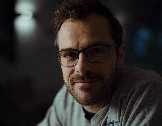 A dimly lit photo of a white man with brown hair, a beard, and glasses, smiling