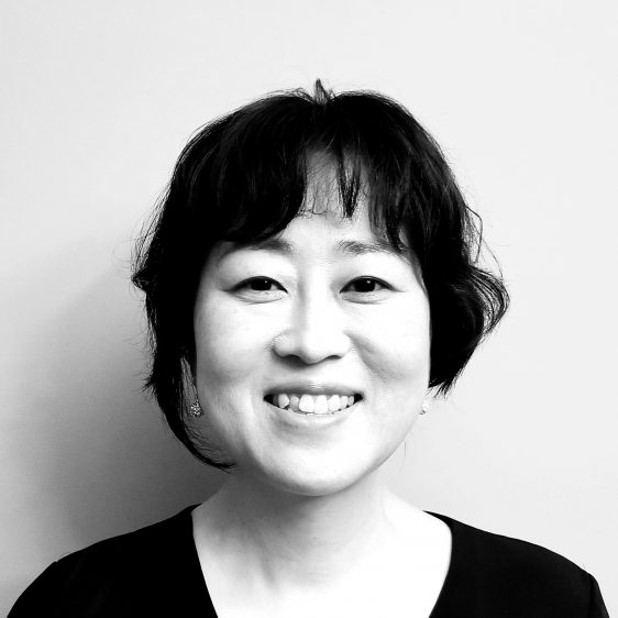 A smiling Korean woman with dark hair framing her face and a black shirt. The photo is black and white.