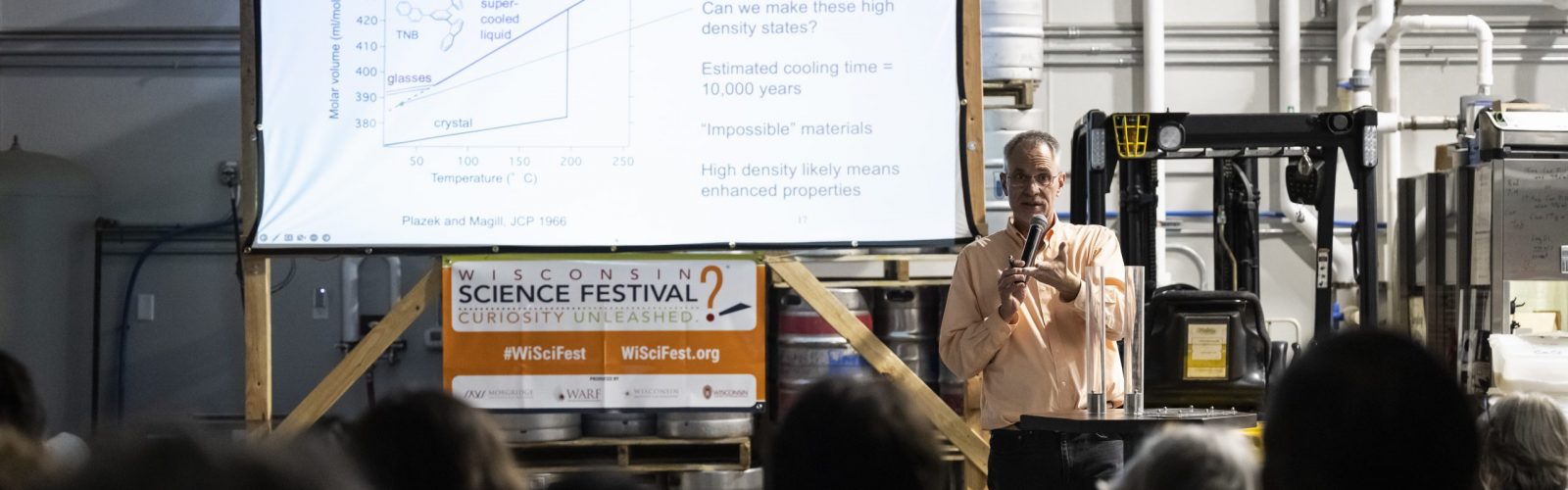 Speaker Mark Ediger stands in front of an elevated projector screen and a banner that reads "Wisconsin Science Festival"