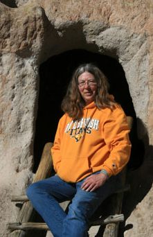 Maureen Muldoon's profile picture. A white woman with long brown hair, wearing an orange sweatshirt, sits in front of an opening in a rock face.