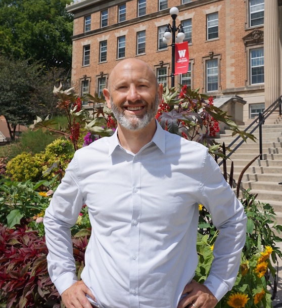 Revel Sims' profile picture. A white man in a white button-down standing with his hands on his hips and smiling in front of a UW Madison building.