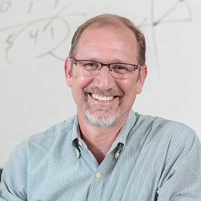 David Williamson Shaffer's profile picture. A white man with glasses and a light green shirt smiles at the camera; behind him is a white board with some markings.