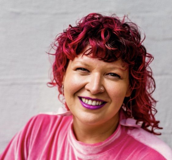 Jess Waggoner's profile picture. A person with pink hair, lipstick, and shirt smiles at the camera against a white background.
