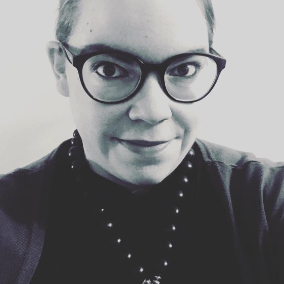 Kate Phelps' profile picture. A person with glasses, a dark shirt, and a necklace smiles at the camera. The photo is black and white.