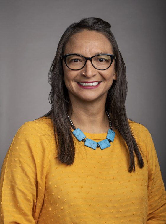Patty Cisneros Prevo's profile picture. A smiling woman with glasses, long brown hair, a yellow shirt and a blue necklace against a grey background.