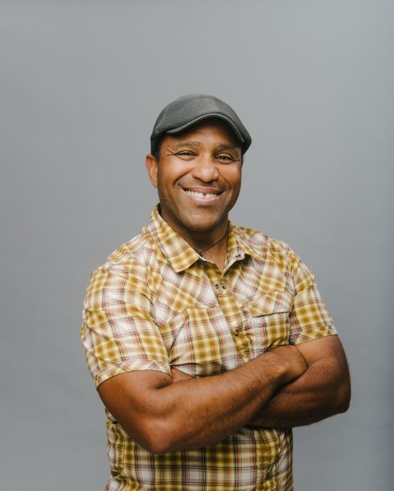 James Mills' profile picture. A Black man in a grey hat and a yellow plaid short-sleeve shirt smiles at the camera against a grey background.