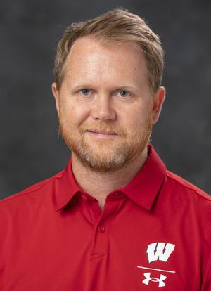 Chad McGehee's headshot. A white man with short blonde hair and a beard, wearing a red Wisconsin athletics shirt, smiles at the camera.